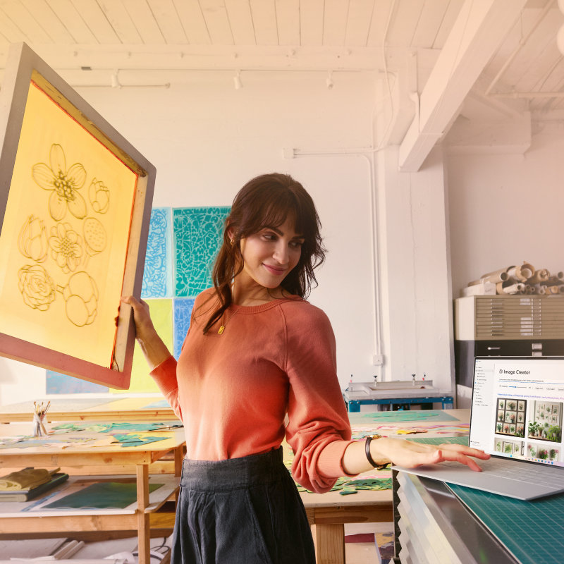 In uno studio d'arte una donna tiene una stampa incorniciata di un fiore nella mano destra, mentre osserva delle immagini su un portatile aperto alla sua sinistra.